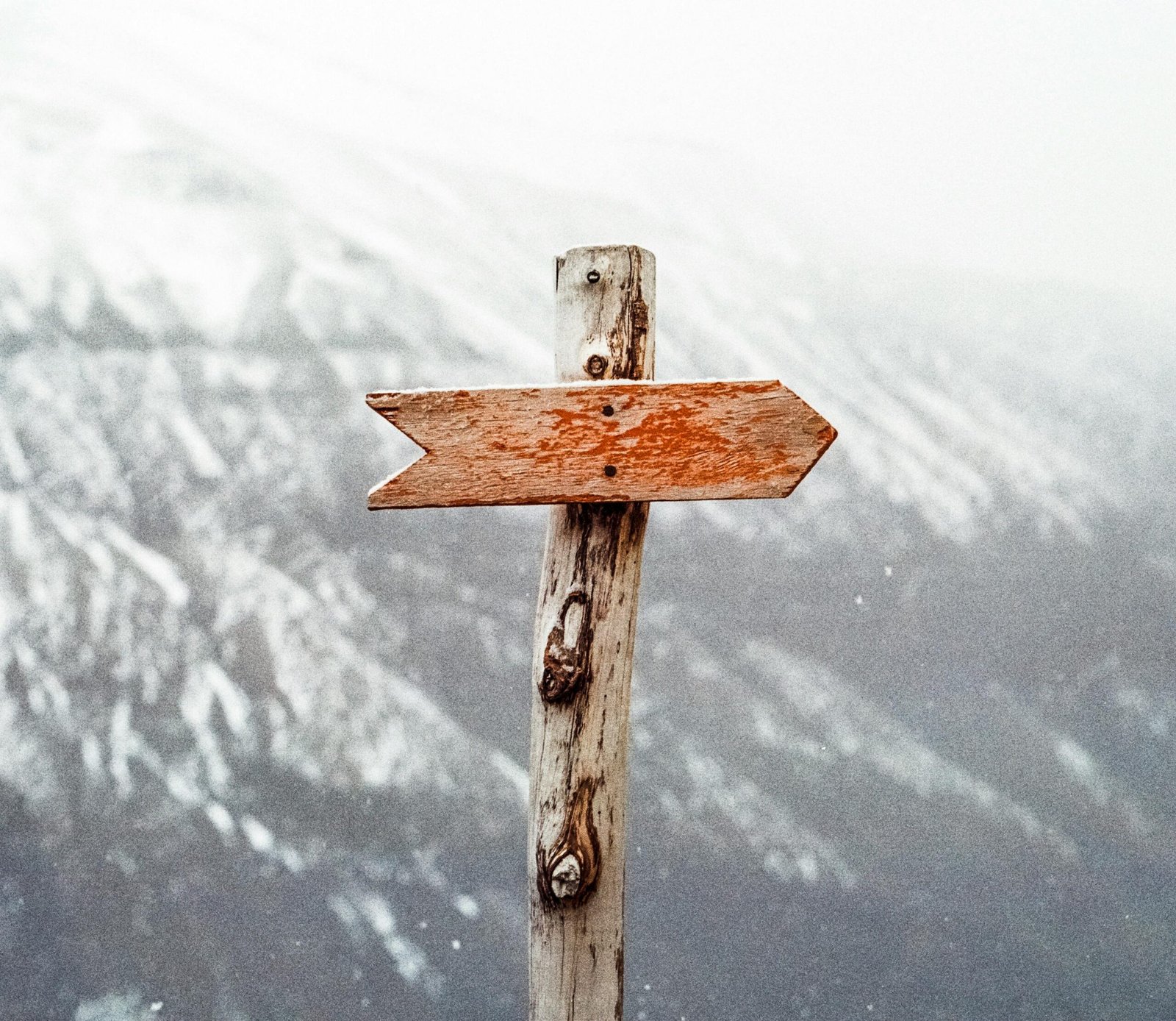 Home A wooden arrow signpost points the way amidst a snowy mountain landscape.